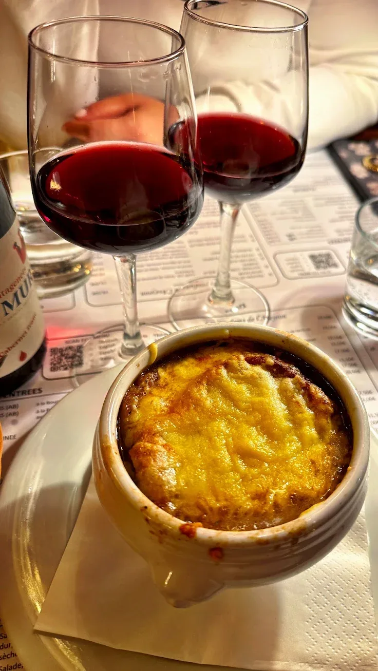 Une soupe à l'oignon gratinée dans son pot en grès, croûte de fromage dorée, avec deux verres de vin rouge